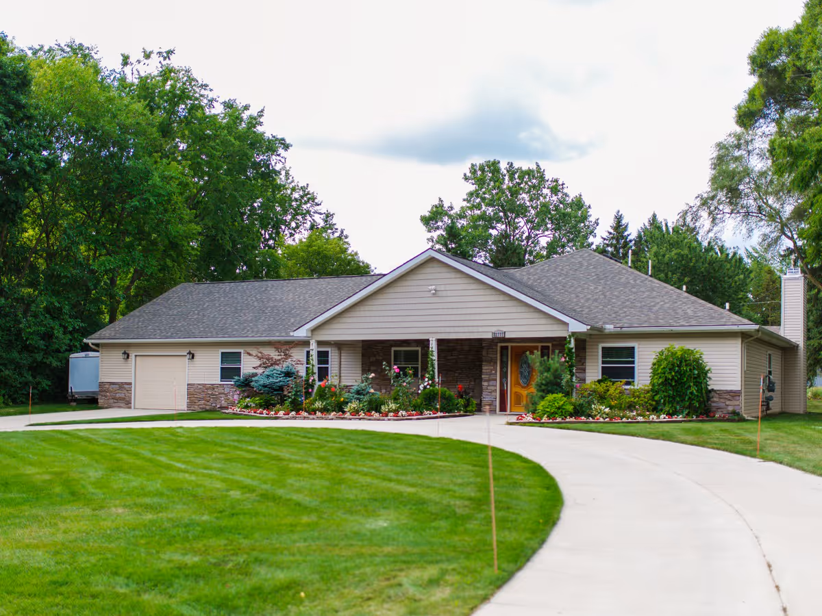 Single-story residential-style building with a gray shingled roof, beige siding, and stone accents. The building has a covered entrance with a wooden door and is surrounded by well-maintained landscaping including bushes, flowers, and trees. A curved concrete driveway leads up to the entrance. The background features tall green trees under a cloudy sky.
