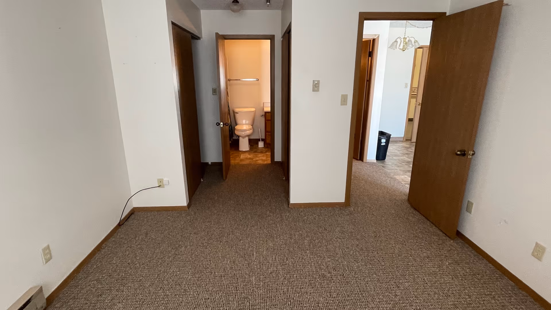 An empty room with beige carpet and white walls. There are two open wooden doors; one leads to a small bathroom with a visible toilet, and the other opens to a hallway with a glimpse of another room. Electrical outlets and a cable are visible on the left wall.