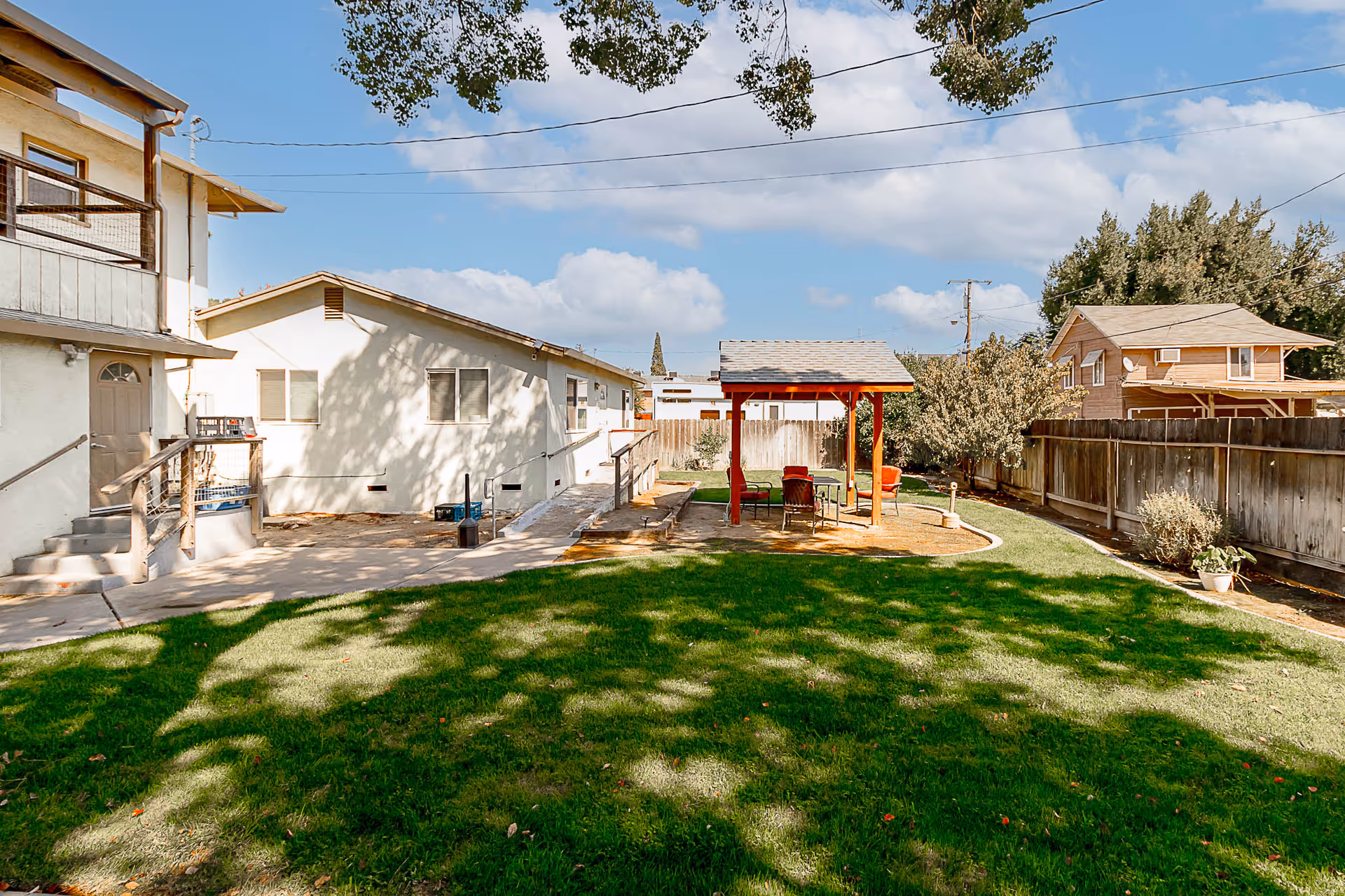 Outdoor area of a senior living facility with a green lawn, a wooden gazebo with chairs and a table underneath, surrounded by a wooden fence and neighboring houses under a partly cloudy sky.