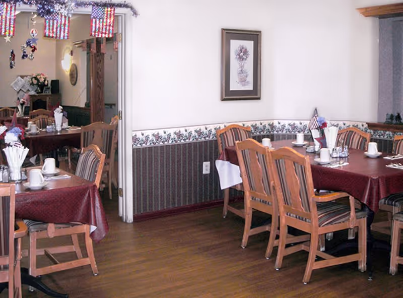 Dining room with wooden chairs and tables covered with maroon tablecloths. Tables are set with white cups, napkins, and utensils. The room has floral wallpaper border and a framed picture on the wall. American flag decorations hang from the ceiling.