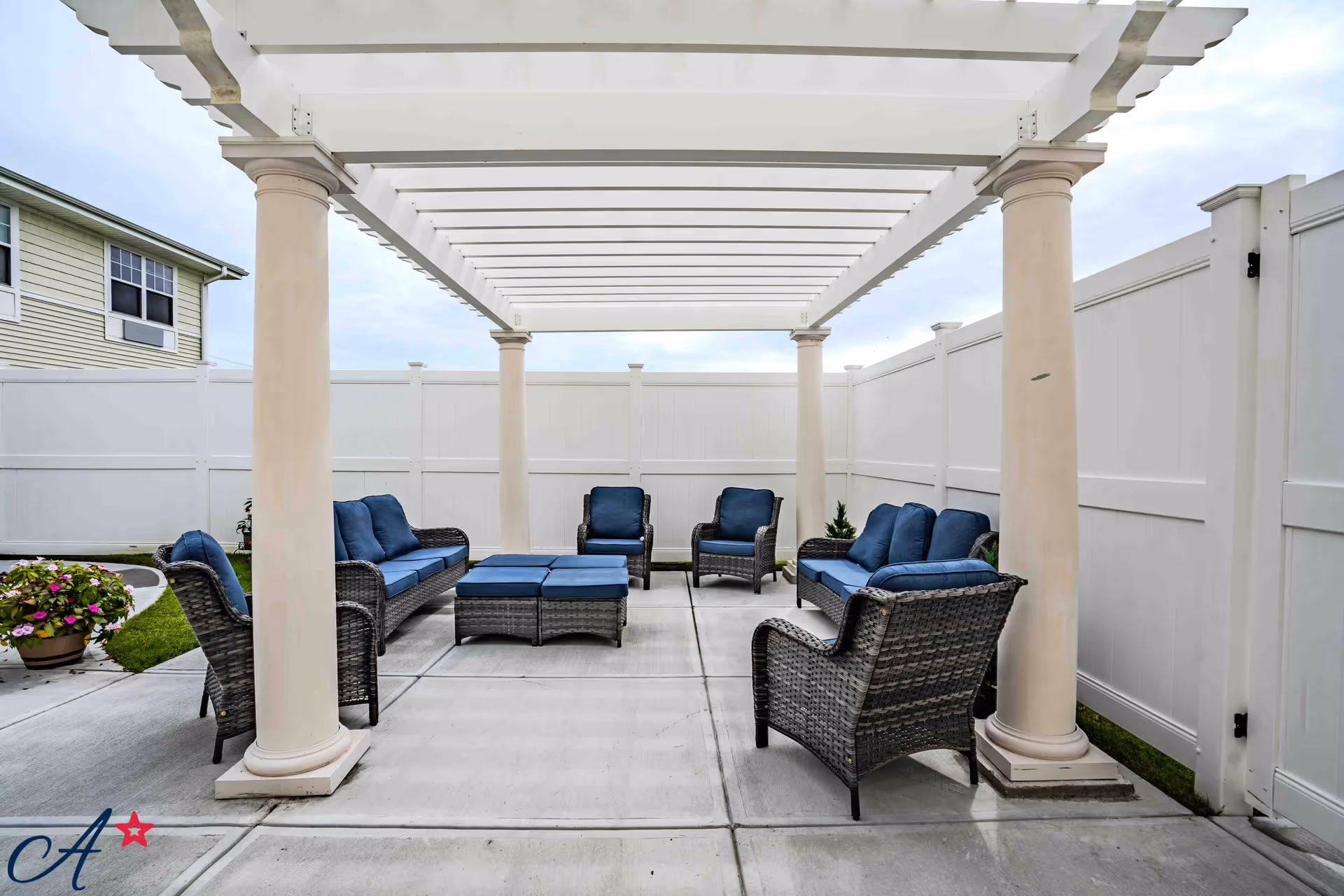 Outdoor seating area with wicker furniture including two sofas, four armchairs, and two ottomans with blue cushions under a white pergola, surrounded by a white privacy fence and a small patch of grass with a potted plant.