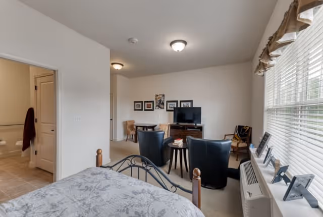 A bright and cozy bedroom with a floral patterned bedspread, two black armchairs facing a TV on a wooden stand, a small round table between the chairs, and a window with blinds and valance letting in natural light. There is a small dining area with two chairs and framed pictures on the wall. A doorway leads to a bathroom with a towel hanging on the wall.
