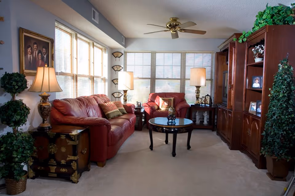 A cozy living room with a red leather sofa and matching armchair, a round glass coffee table, two table lamps, a ceiling fan, large windows with blinds, wooden shelving unit with framed photos, and decorative plants.