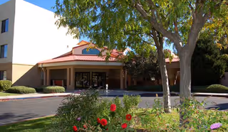 Covered front entrance of a senior living building with a circular driveway, trees, and flowering shrubs in the foreground.