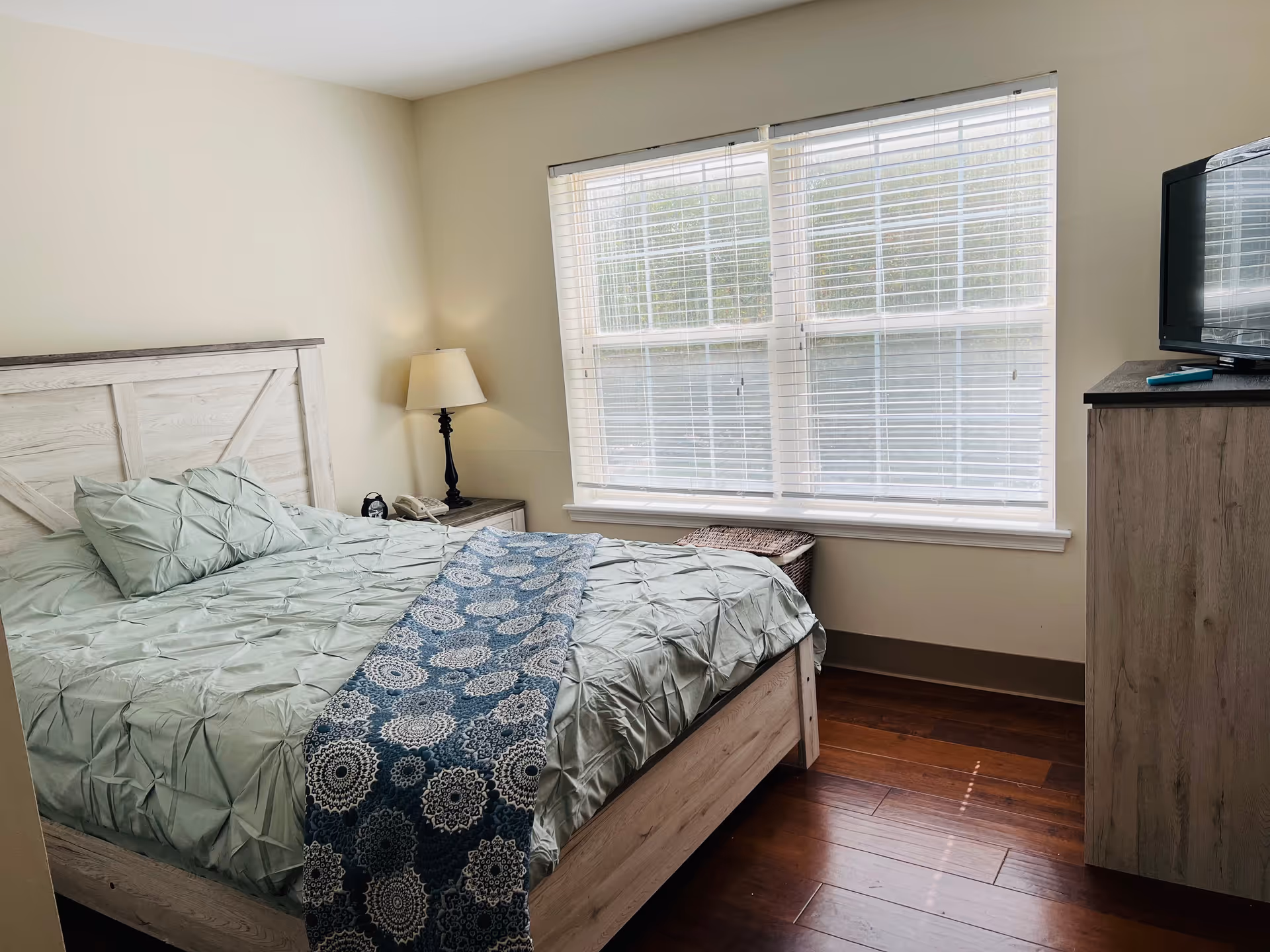 A bedroom with a wooden bed frame and light green bedding, a blue patterned runner on the bed, a nightstand with a lamp and a phone, a large window with white blinds, a wicker basket under the window, and a wooden dresser with a TV on top.