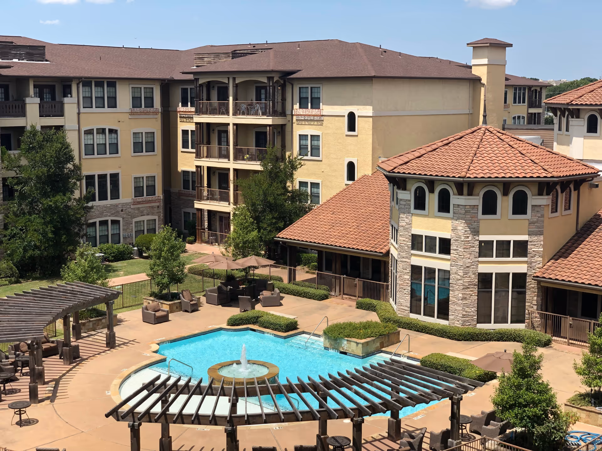 Outdoor view of a senior living facility featuring a swimming pool with a central fountain, surrounded by patio seating areas with umbrellas and pergolas. The background shows a multi-story building with balconies and a tiled roof under a clear blue sky.