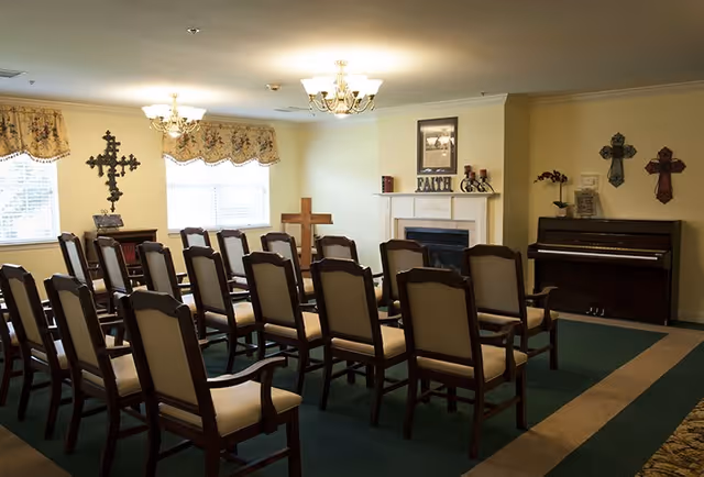 A small chapel or prayer room with rows of wooden chairs with beige cushions facing a fireplace and a piano. The room has several decorative crosses on the walls, floral valances on the windows, and a green carpet with a beige border.