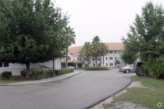 Exterior view of a senior living campus with a driveway, trees, and parked cars.