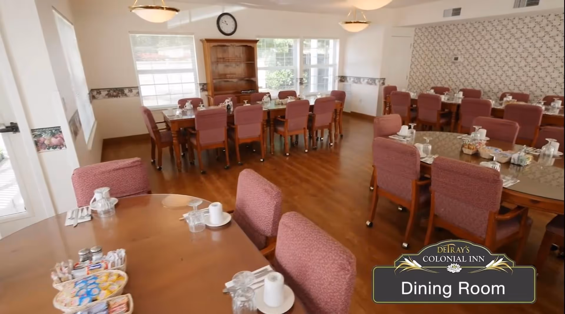 A bright dining room with multiple wooden tables and cushioned chairs arranged neatly. The tables are set with glasses, cups, and condiments. Large windows allow natural light to fill the room, and there is a wooden cabinet against the far wall. The floor is wooden, and the walls are decorated with a floral wallpaper border and a patterned accent wall.