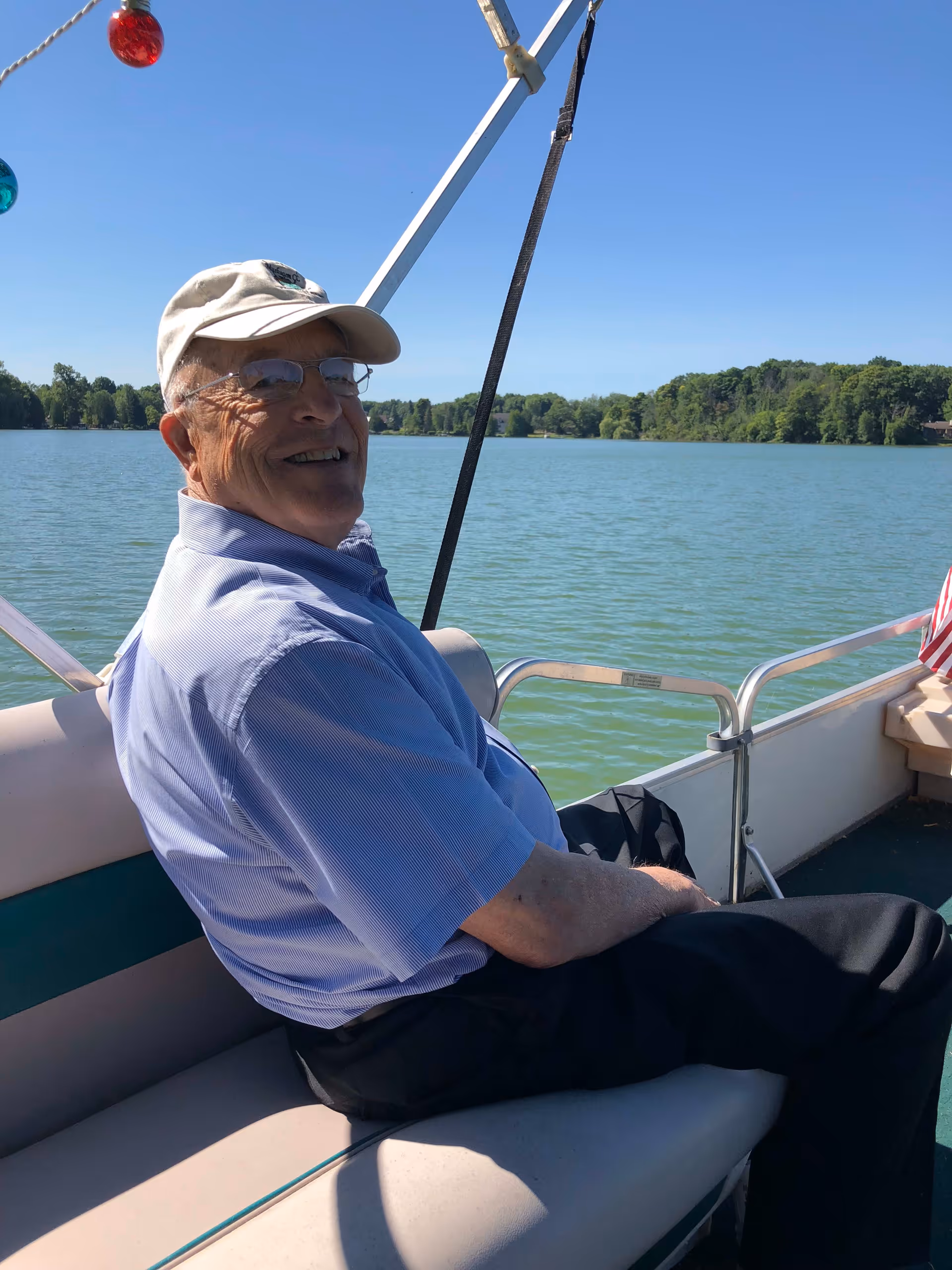 An elderly man wearing a light blue shirt, black pants, glasses, and a white cap is sitting and smiling on a boat with a lake and green trees in the background under a clear blue sky.