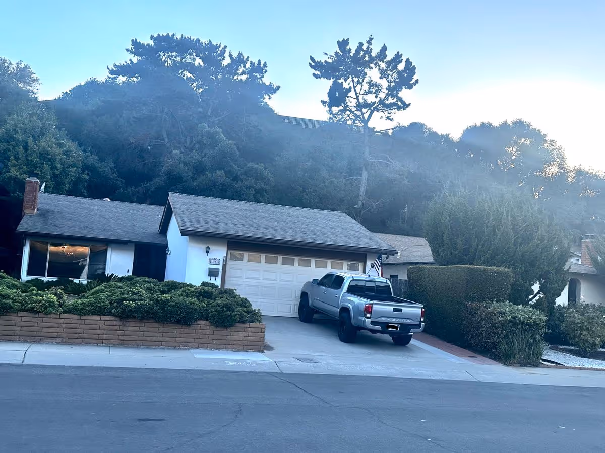 Single-story house with an attached garage and a pickup truck parked in the driveway, set against a wooded hillside.