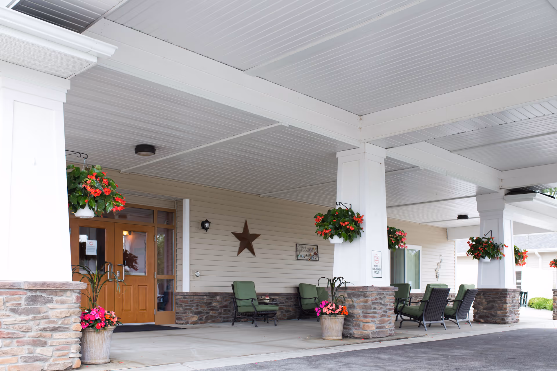 Covered entrance area of a senior living facility with stone and white pillars, hanging flower baskets, potted plants, green cushioned chairs, and a wooden double door entrance with a star decoration on the wall.