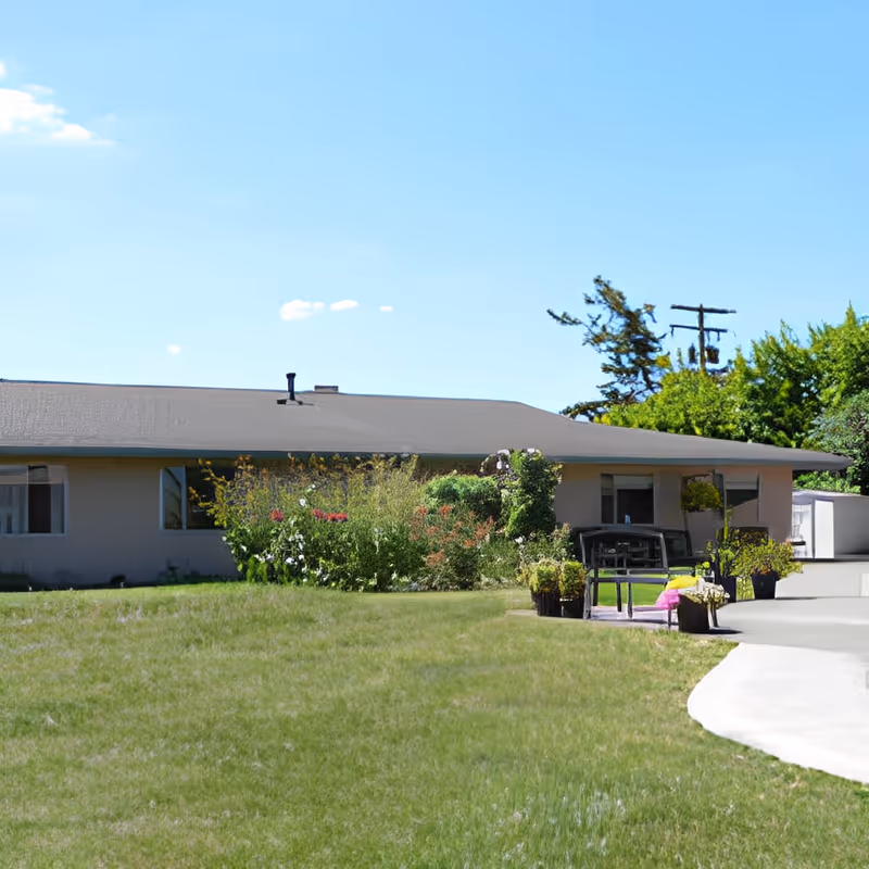Single-story building with a gray roof surrounded by green grass and various plants and shrubs. There is a small patio area with outdoor furniture and potted plants. The sky is clear and blue with a few small clouds.