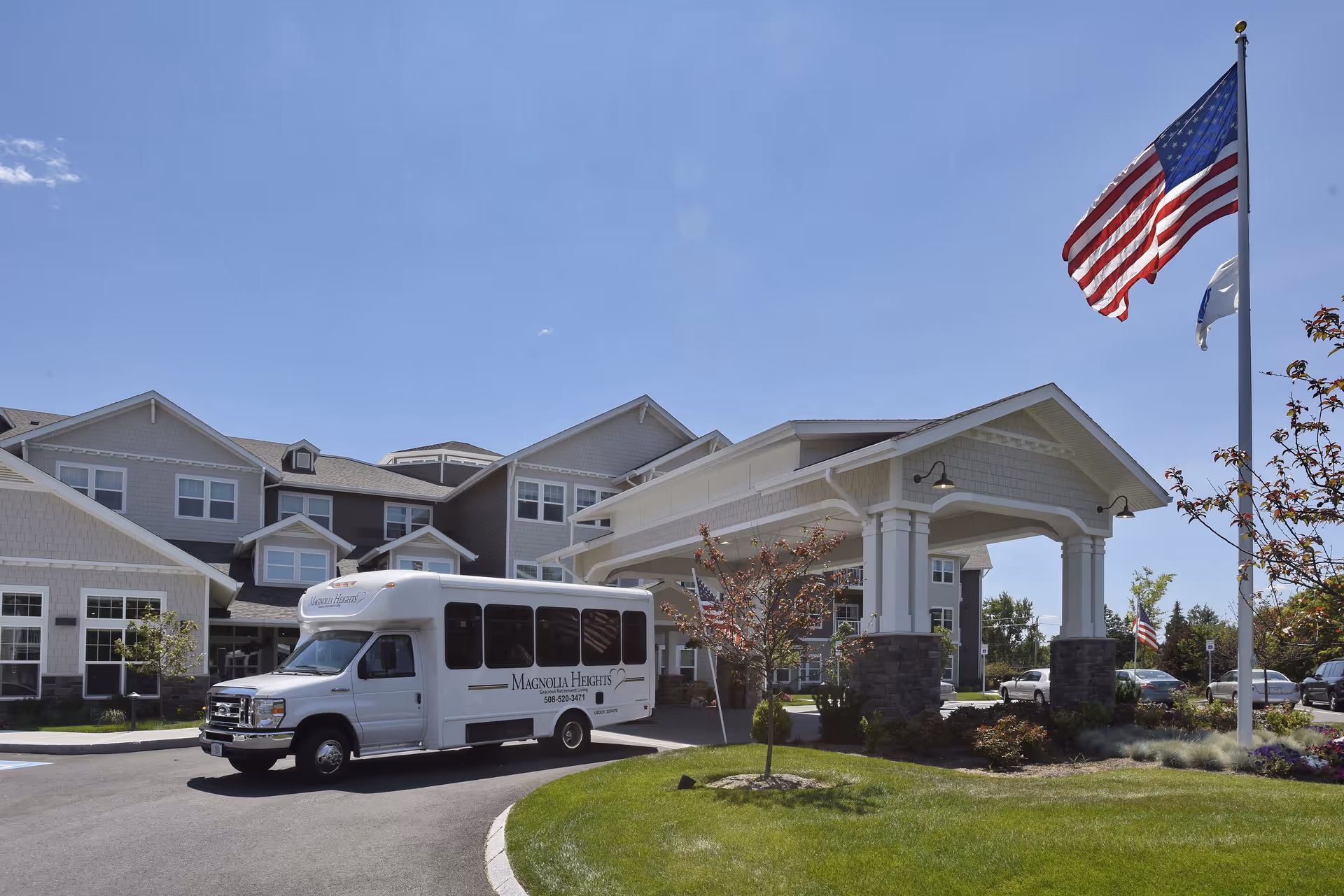 Exterior view of Magnolia Heights Gracious Retirement Living facility on a sunny day with a white shuttle bus parked near the entrance. The building has multiple windows and a covered drop-off area. An American flag and another flag are flying on flagpoles near the entrance, with green grass and small trees in the foreground.