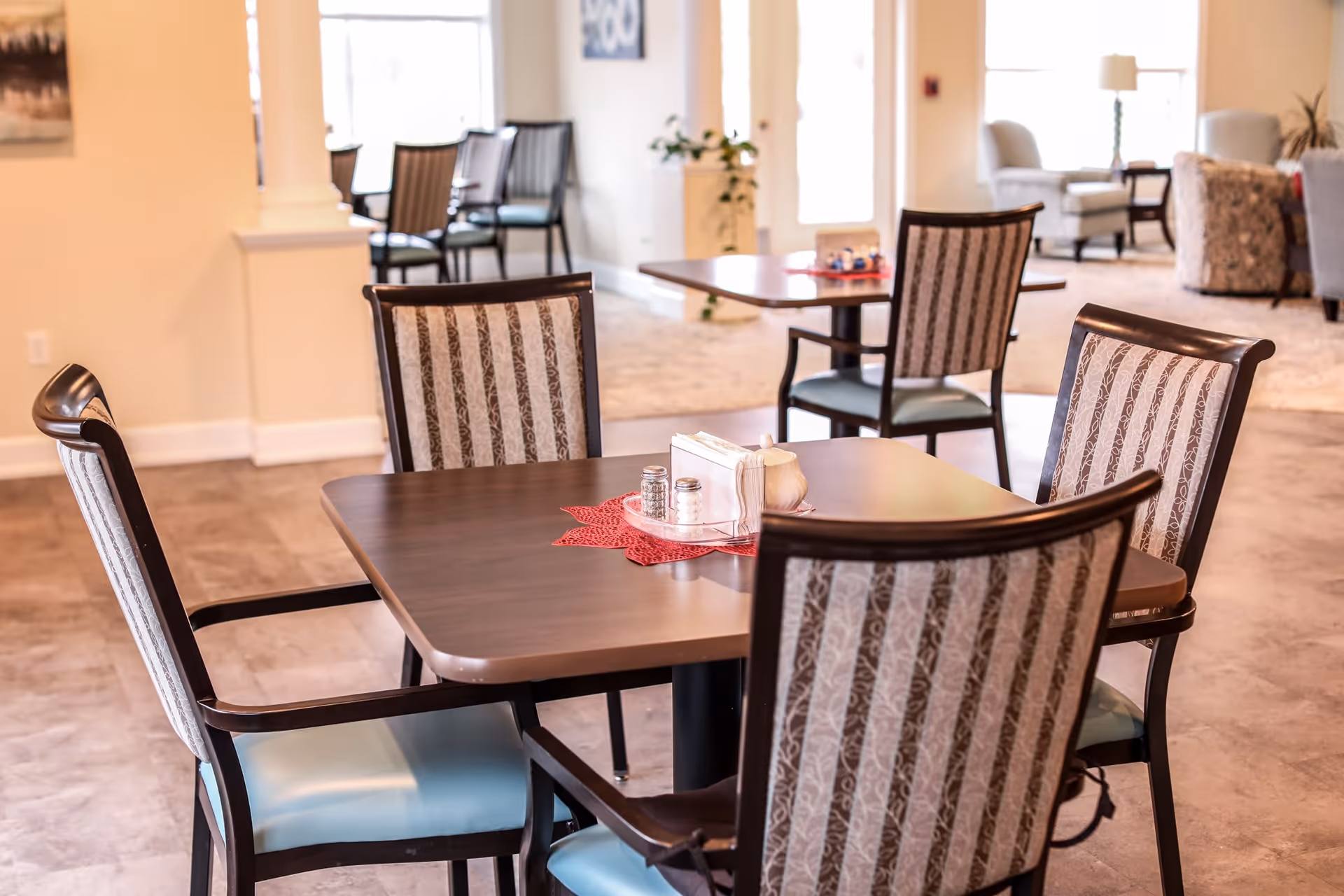A dining area in an assisted living facility with a wooden table and four chairs with patterned upholstery. The table has a red placemat, salt and pepper shakers, napkins, and a small container. In the background, there are more tables and chairs, a lamp, and some armchairs in a well-lit room with large windows.