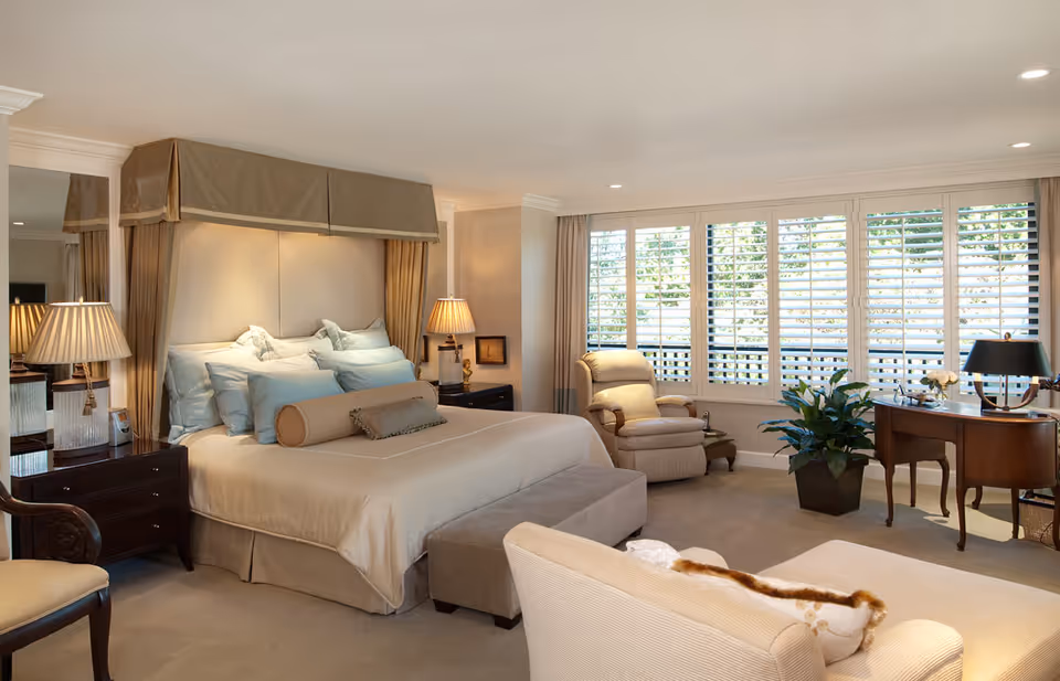 A spacious and elegantly decorated bedroom featuring a large bed with multiple pillows and a canopy headboard. The room includes two bedside tables with lamps, a comfortable armchair near large windows with white plantation shutters, a wooden desk with a lamp, and a beige sofa in the foreground. The room is well-lit with natural light and recessed ceiling lights.