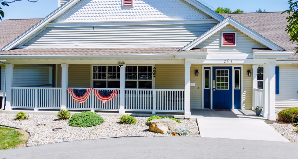 Front exterior view of a single-story senior living facility building with a covered porch, white railing, and blue double doors. The porch is decorated with red, white, and blue bunting. There are small bushes and rocks in the landscaped area in front of the building.