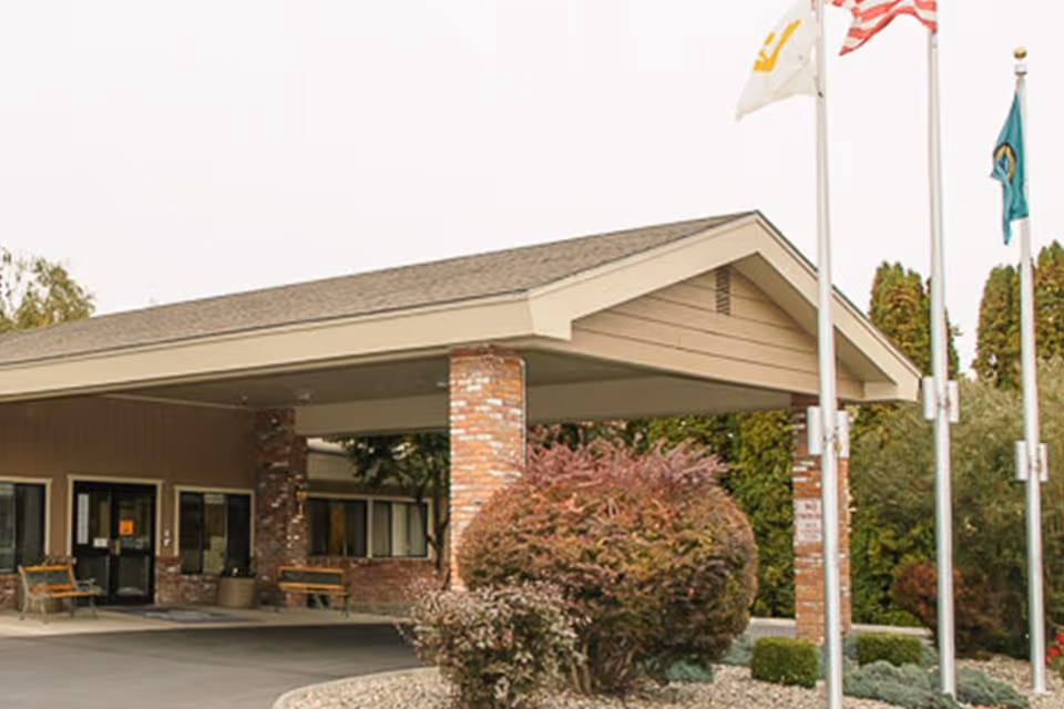 Entrance of a single-story building with a covered driveway supported by brick pillars. There are benches near the entrance doors and three flagpoles with flags flying. Shrubs and trees surround the area.