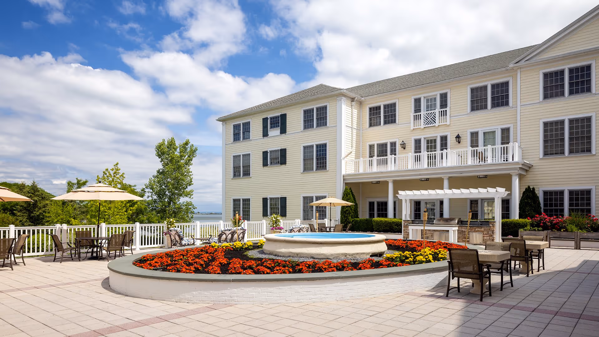 Outdoor patio area at Atria on Roslyn Harbor featuring a circular flower bed with vibrant red and yellow flowers surrounding a small fountain. Several tables with chairs and umbrellas are arranged around the patio. The background shows a three-story light yellow building with multiple windows and a balcony, under a partly cloudy blue sky.