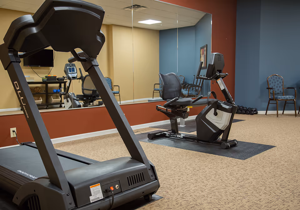 Indoor fitness room with a treadmill and a recumbent exercise bike on carpeted floor. The room has a large mirror on one wall, several chairs along the walls, and a rack with dumbbells in the corner. The walls are painted in blue, beige, and red tones.