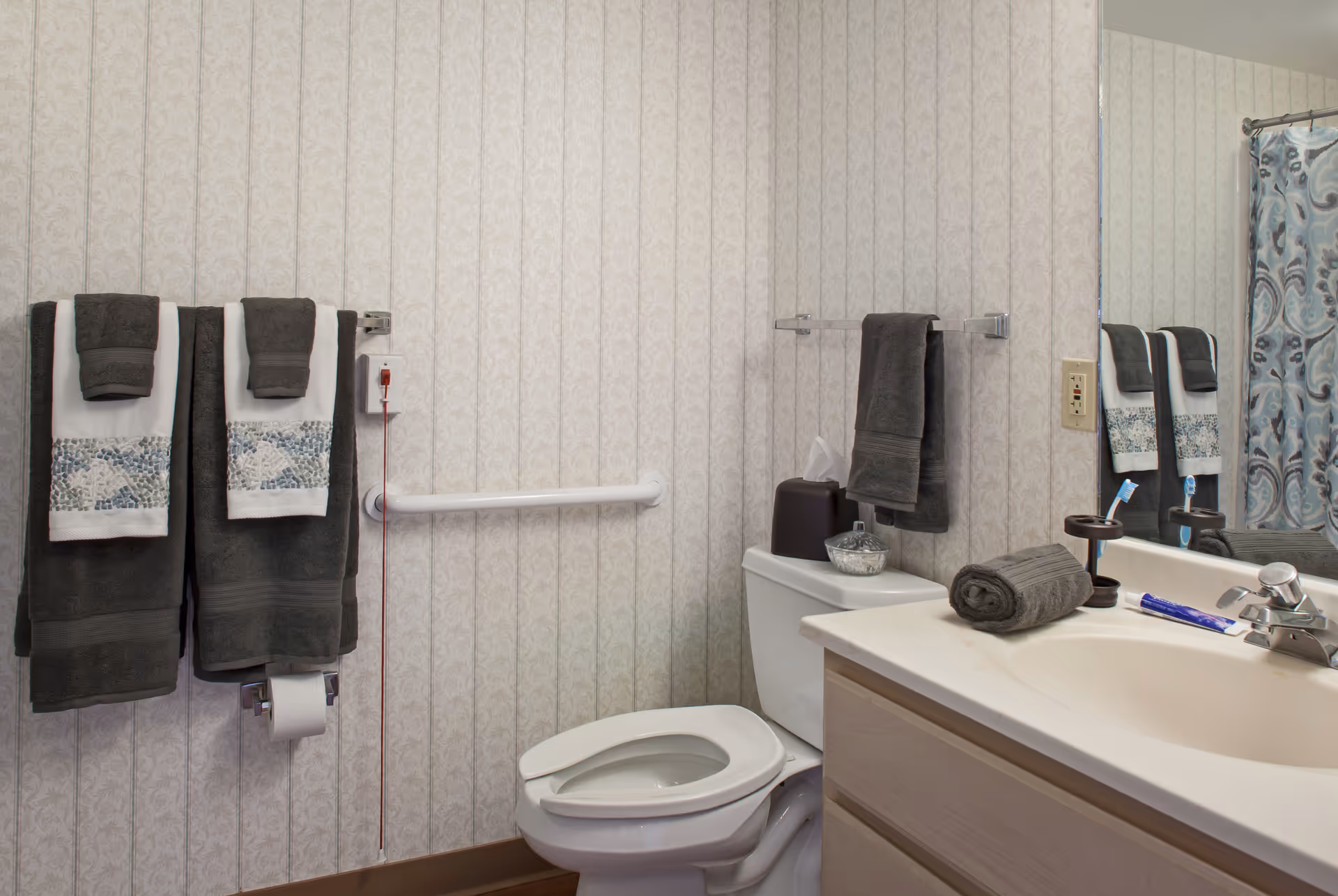A bathroom with a toilet, a sink with a faucet, and a large mirror. The toilet has a tissue box and a small container on top. There are dark gray towels hanging on towel racks and a patterned shower curtain visible on the right side. The walls have a light patterned wallpaper, and there is a grab bar next to the toilet.