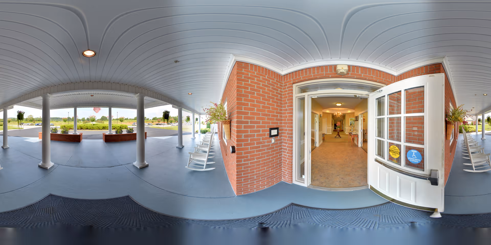 Covered entrance area of a senior living facility with white rocking chairs lined up along the brick wall. The entrance door is open, showing a hallway inside with carpeted flooring and warm lighting. Outside, there are columns supporting the roof and planters with greenery. The view extends to a parking lot and landscaped grounds in the background.