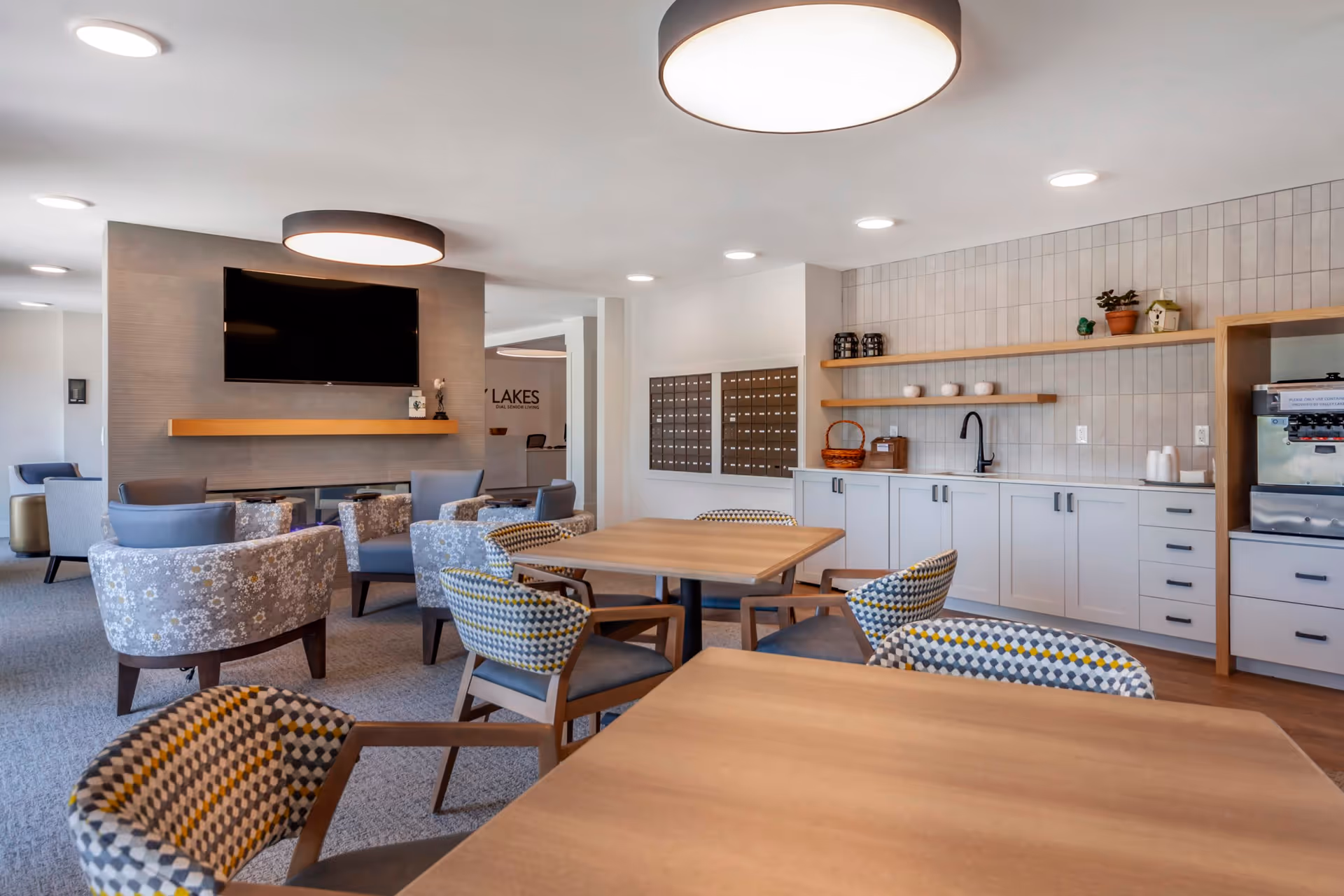 A modern and cozy common area in an assisted living facility featuring several wooden tables with patterned chairs, a seating area with floral upholstered armchairs, a wall-mounted flat screen TV above a wooden shelf, and a kitchenette with white cabinets, open wooden shelves holding decorative items, and a coffee machine.