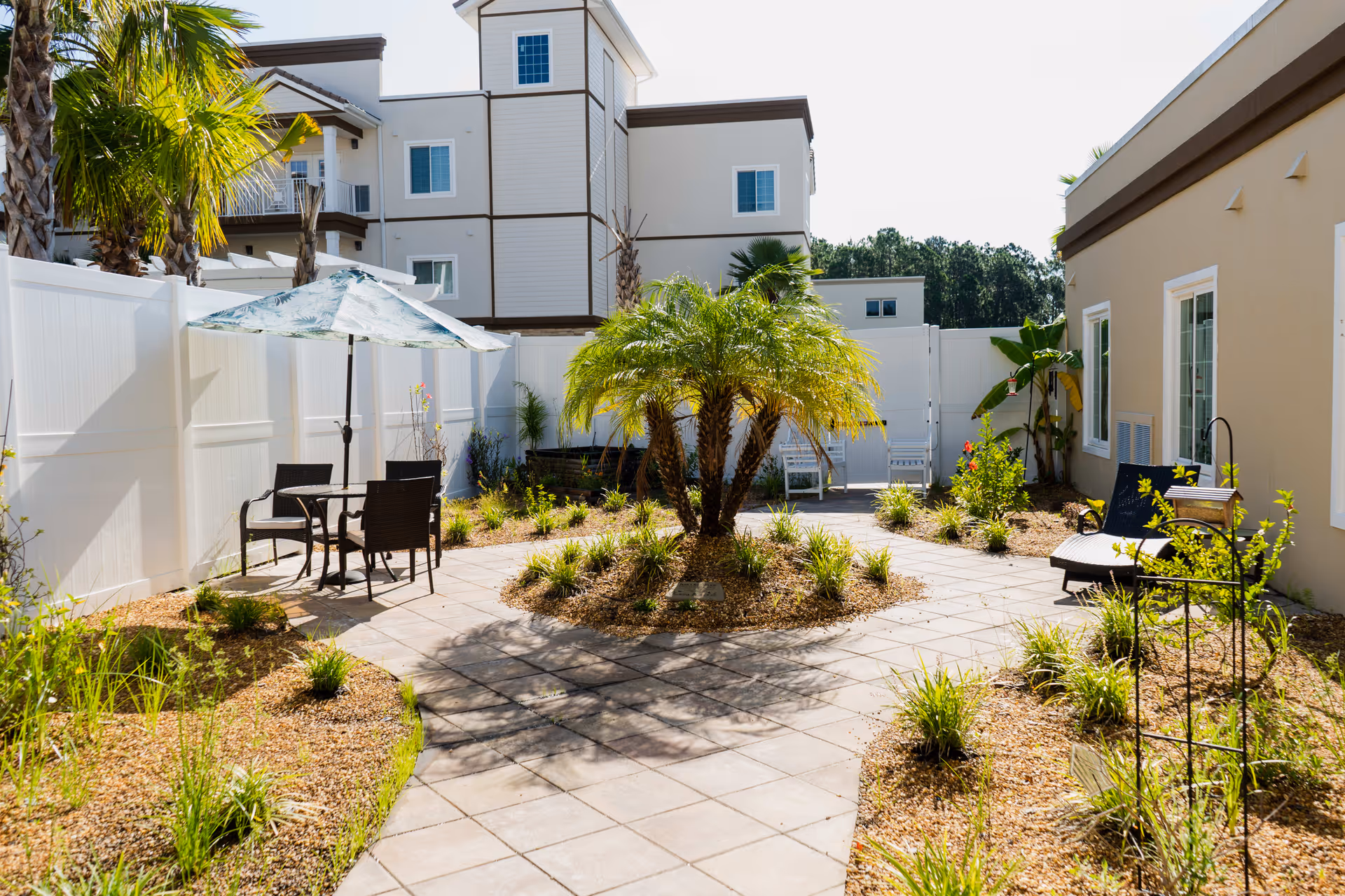 Outdoor courtyard area with a paved walkway, a central palm tree surrounded by small plants, patio table with umbrella and chairs on the left, and lounge chairs with plants on the right. The area is enclosed by white fencing and adjacent to a beige building with windows.