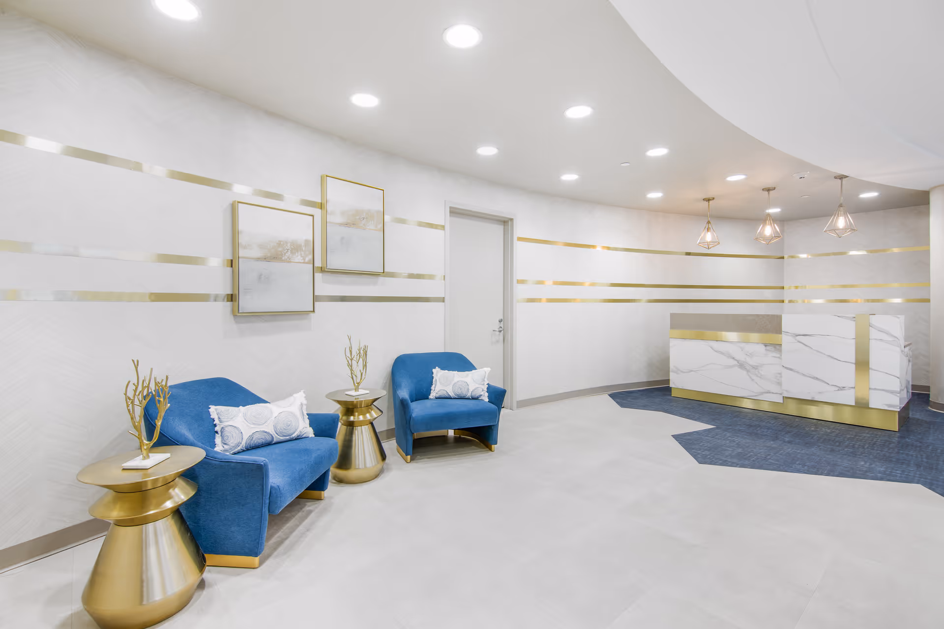 A modern and elegant reception area with two blue armchairs, each with a decorative pillow, and two gold side tables with decorative branches. The walls are white with horizontal gold stripes and two framed abstract artworks. The reception desk is white with a marble pattern and gold accents, and three pendant lights hang above it. The floor is a combination of light tiles and dark blue carpet.