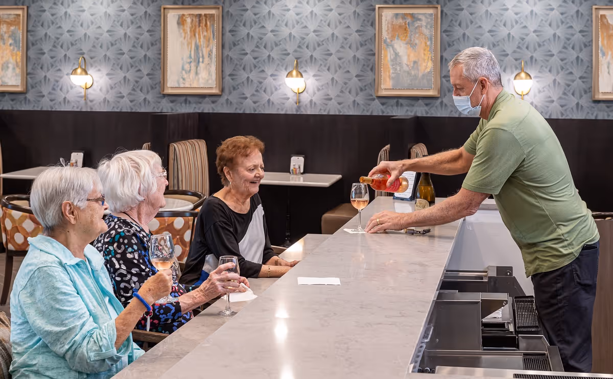 Three elderly women sitting at a bar counter in a dining area, each holding a glass of rosé wine, while a man wearing a face mask pours wine into a glass. The background features patterned wallpaper, framed artwork, and wall-mounted lights.