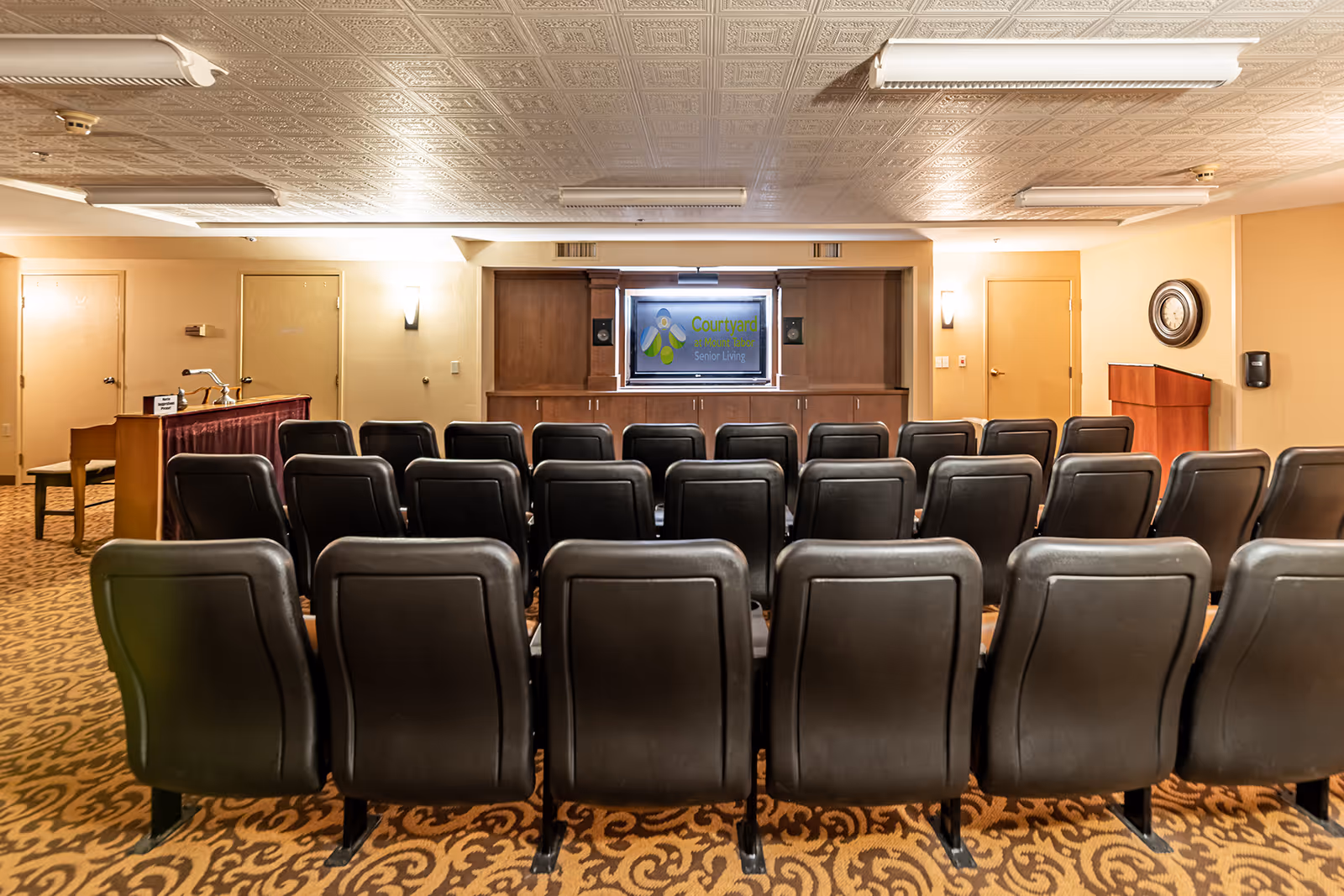 A small auditorium or meeting room with several rows of black chairs facing a wooden cabinet with a screen displaying the Courtyard at Mount Tabor Senior Living logo. The room has beige walls, a patterned carpet, a clock on the right wall, and a podium near the right side.