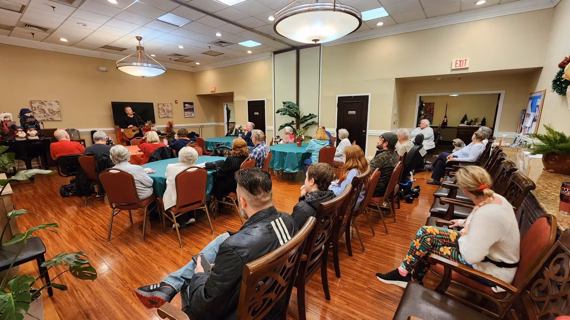 A group of people seated in a community room with wooden floors and beige walls, attentively watching a man playing guitar near a piano. The room has round tables with green tablecloths and chairs arranged in rows. There are plants and holiday decorations around the room.