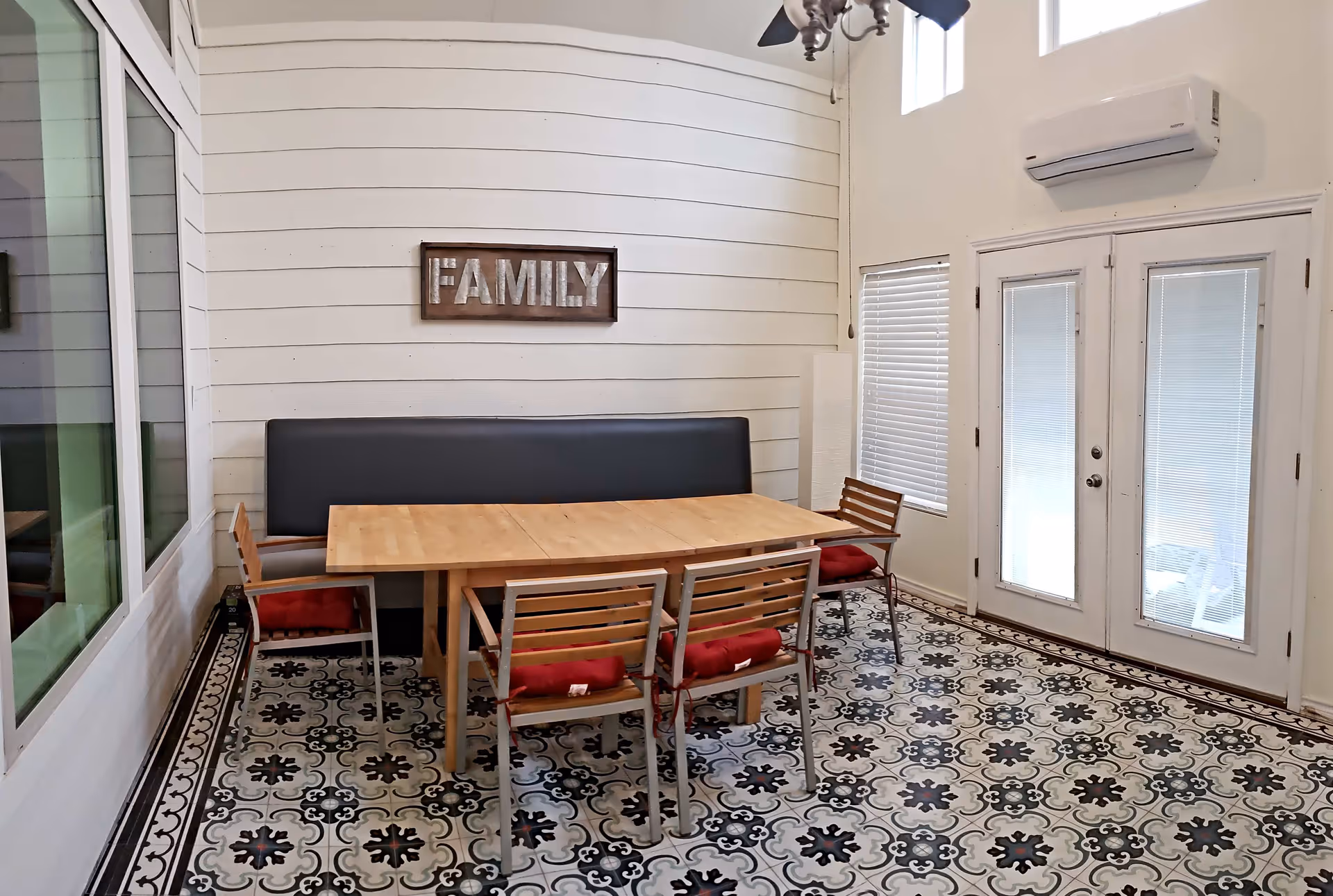 A dining area with a wooden table surrounded by four wooden chairs with red cushions and a black cushioned bench against a white shiplap wall. Above the bench is a wooden sign with the word 'FAMILY'. The floor has a decorative black, white, and gray patterned tile. There are two windows with blinds and a set of white French doors with glass panels. A ceiling fan is partially visible at the top.