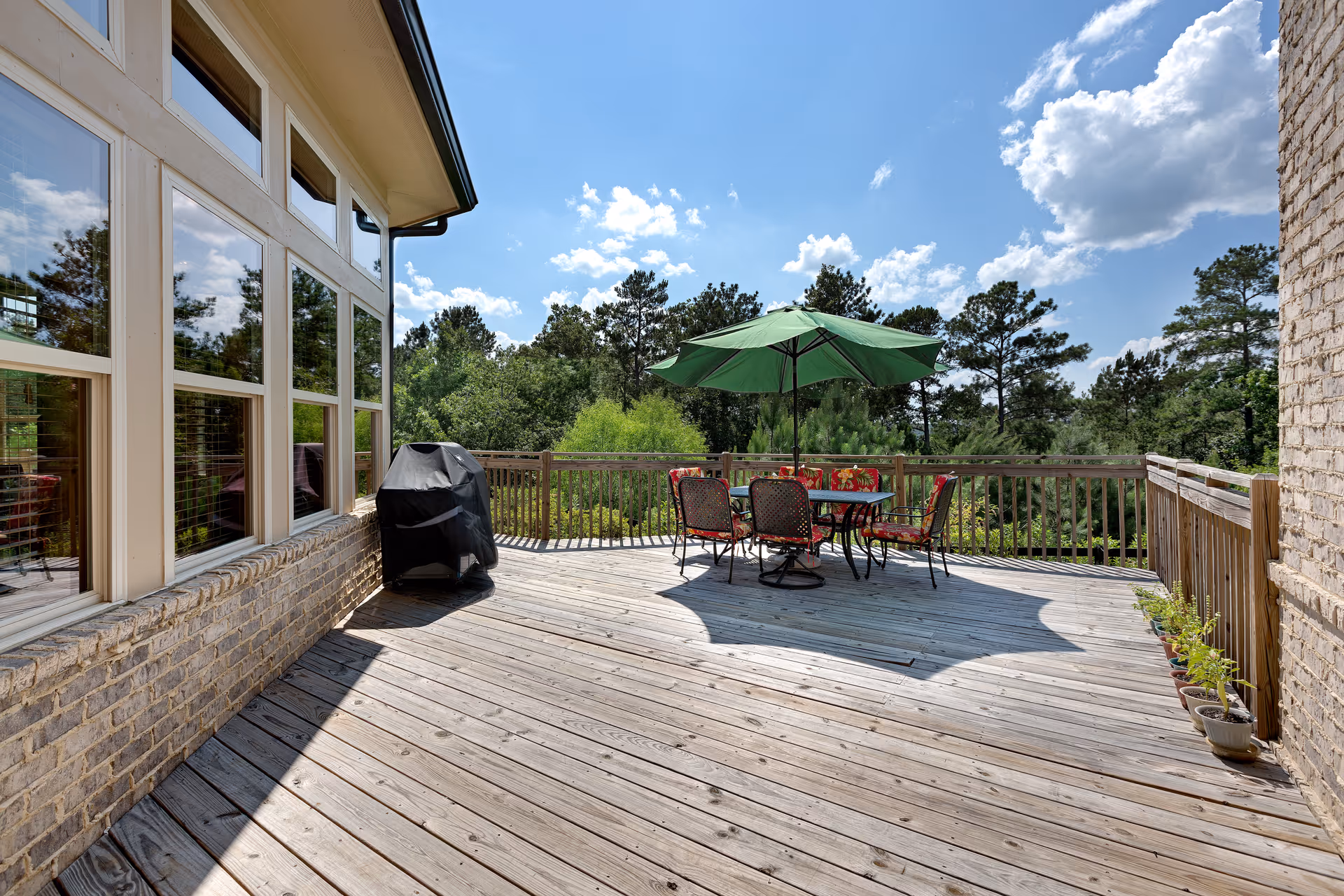 A spacious wooden deck with a table and four chairs under a green patio umbrella. There is a covered grill near the windows of a brick building. The deck overlooks a green wooded area under a blue sky with some clouds.