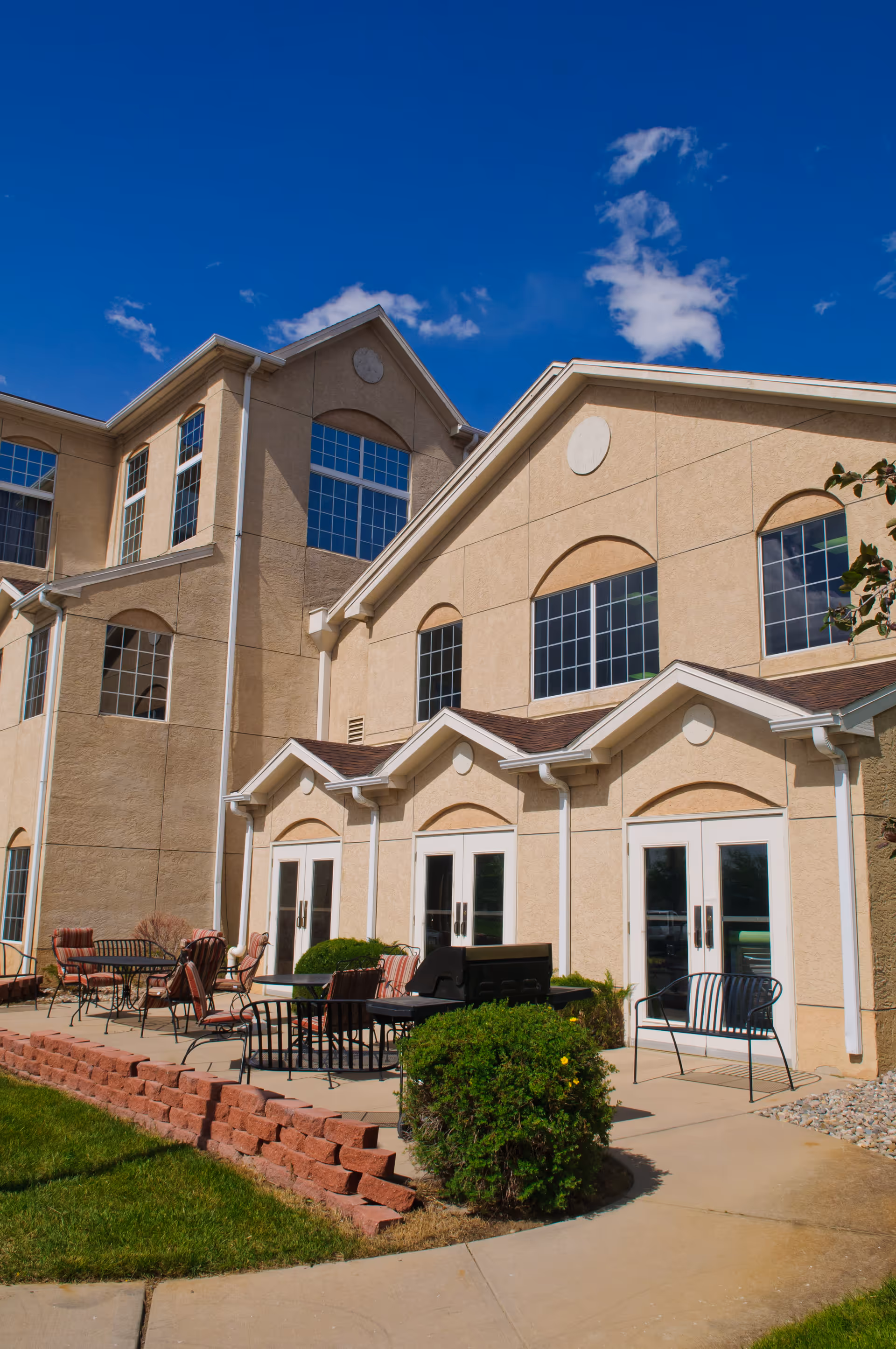 Multi-story beige building exterior with arched windows, glass doors and a patio area with tables and chairs under a blue sky.