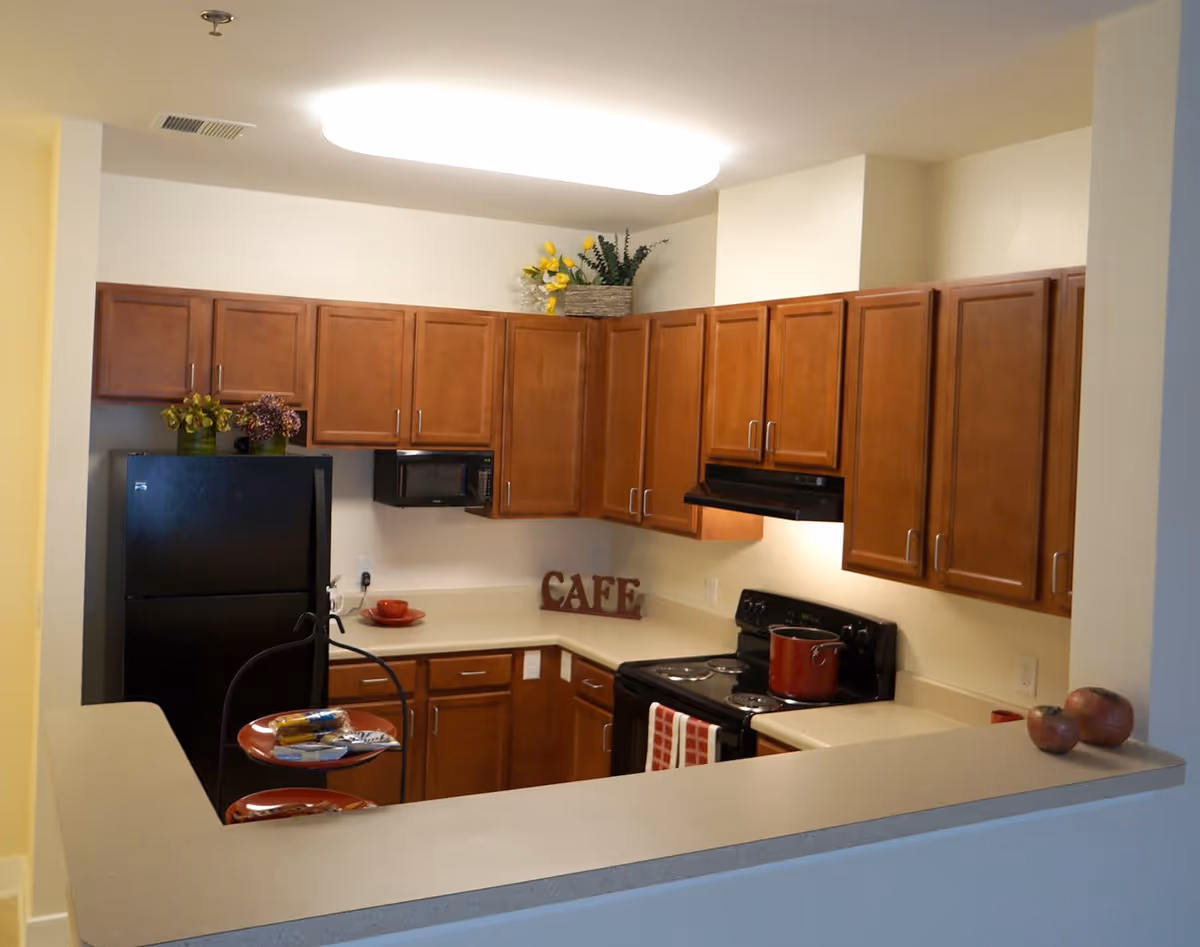 A kitchen with wooden cabinets, a black refrigerator, a black microwave, and a black stove with a red pot on it. There are decorative plants on top of the cabinets and a sign that says 'CAFE' on the countertop. A two-tiered tray with snacks is on the counter in the foreground.