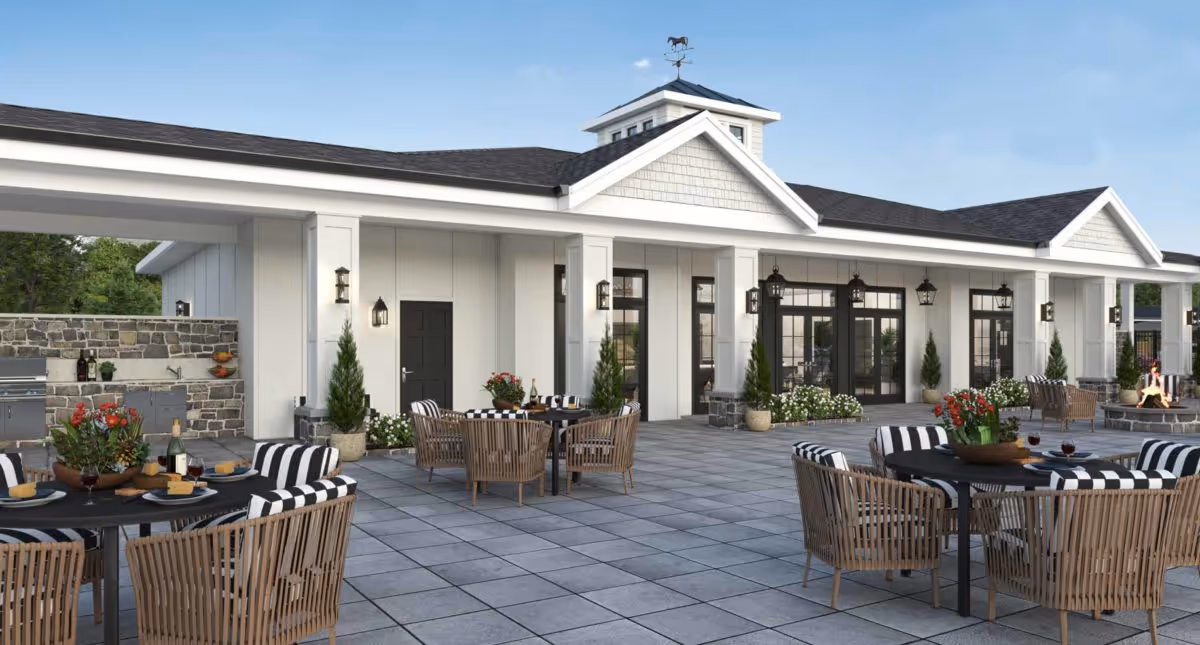 Outdoor patio area of a senior living facility with multiple round tables surrounded by wicker chairs with black and white striped cushions. The patio is paved with large square tiles and features a built-in stone grill and countertop area. The building has white walls, black doors, and large windows with black frames. There are potted plants and flowers decorating the space, and a clear blue sky is visible above.