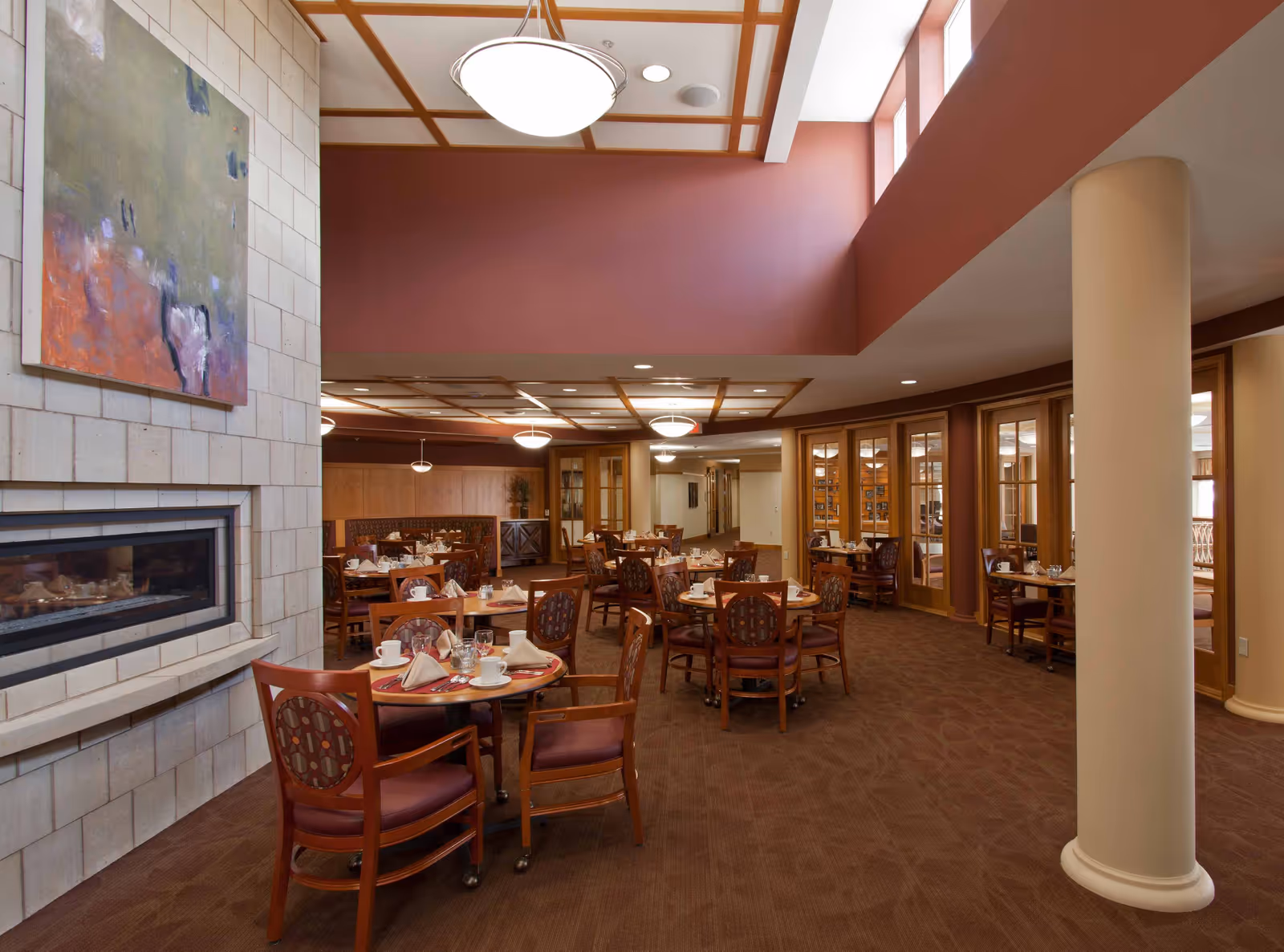 A spacious dining room with multiple round wooden tables set with white napkins, cups, and glasses. The room features a modern fireplace on the left wall with an abstract painting above it. The ceiling has wooden beams and recessed lighting, and there are large windows near the ceiling allowing natural light to enter. The carpet is brown, and the walls are painted in warm tones.