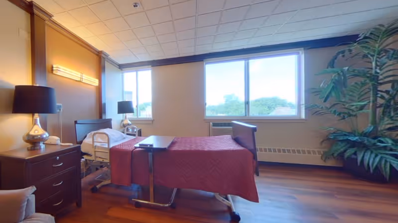 A well-lit bedroom in a senior living facility featuring a hospital-style bed with a maroon quilt, a bedside table with a lamp, a large window with a view of trees outside, and a large potted plant in the corner.