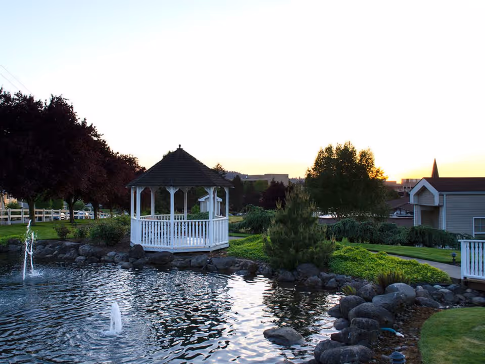 A serene outdoor scene at sunset featuring a white gazebo beside a pond with two small water fountains. The area is surrounded by green grass, rocks, trees, and shrubs, with buildings visible in the background.