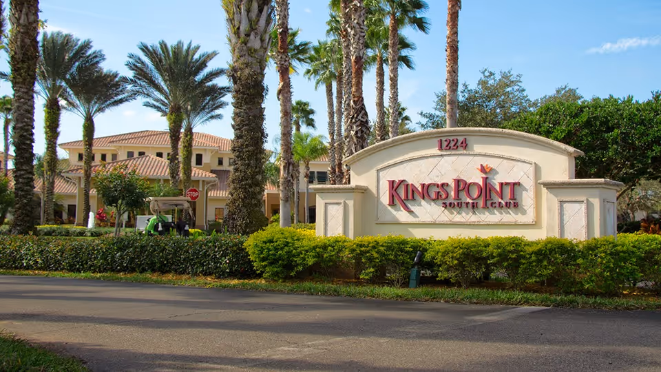 Large stone entrance sign reading 'Kings Point Suncoast' with palm trees and a Mediterranean-style building behind it.