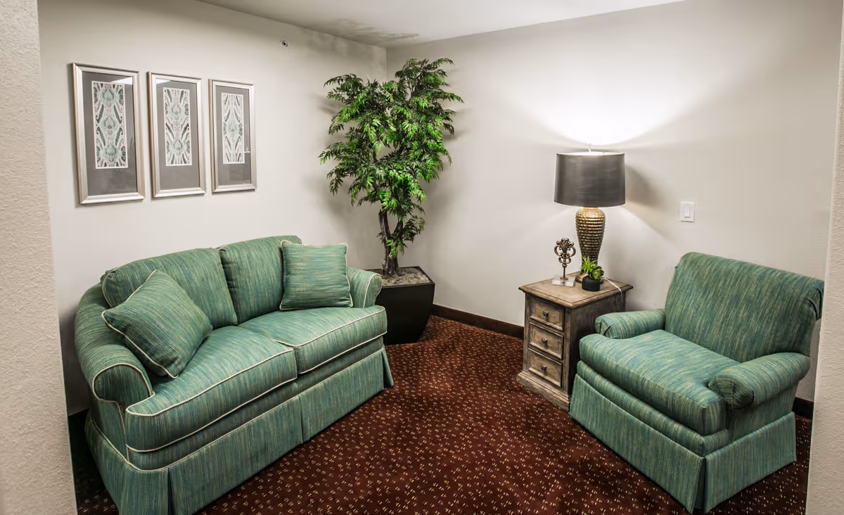 A cozy sitting area with a green upholstered sofa and matching armchair, a wooden side table with a lamp and decorative items, a potted plant in the corner, and three framed artworks on the wall.