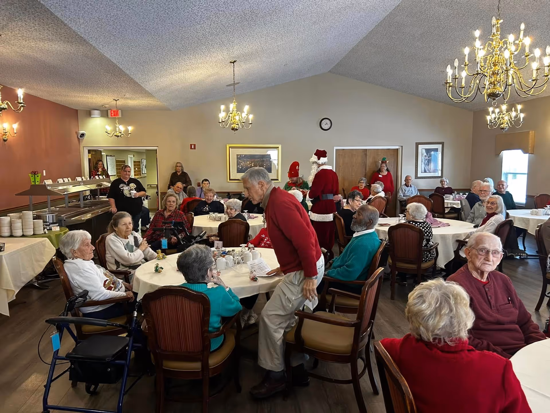 A group of elderly people gathered in a dining room with round tables and chairs. Some are seated while others are standing and interacting. The room is decorated with chandeliers and has a buffet area with plates and bowls. Two people dressed as Santa Claus and an elf are present, suggesting a holiday event.