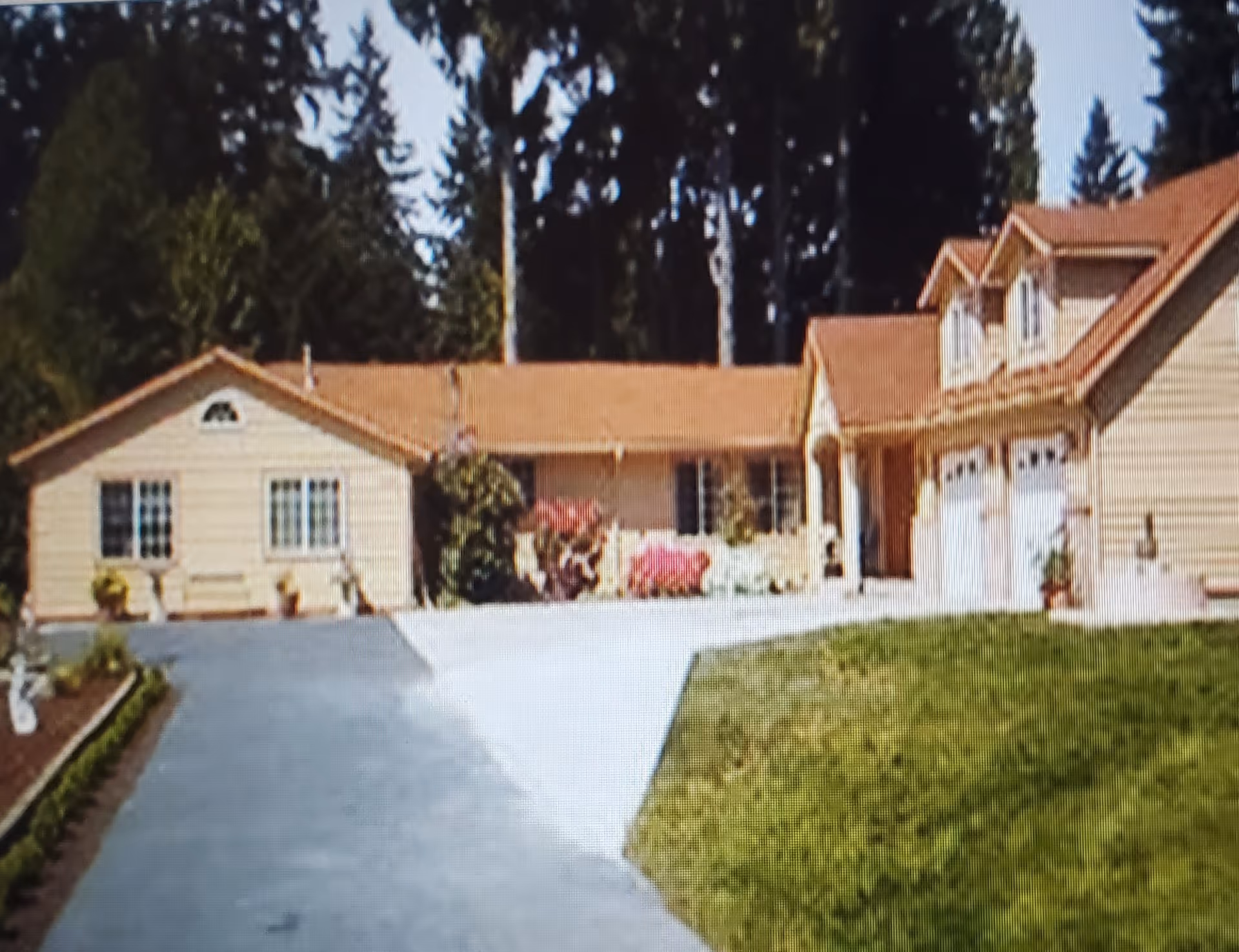 Exterior view of a single-story residential building with beige siding and a brown roof, featuring a driveway, green lawn, and some bushes and flowers in front. Tall trees are visible in the background.