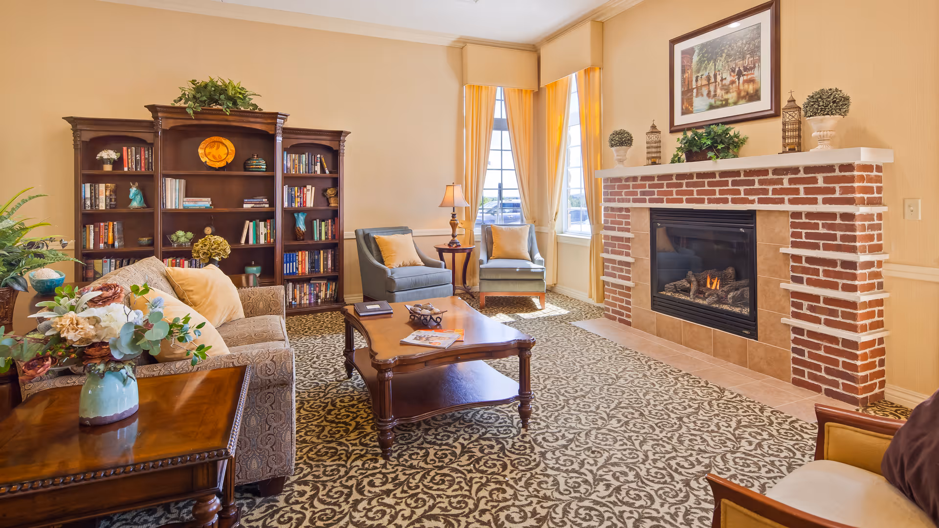 A cozy living room with a patterned carpet, a brick fireplace with a fire burning, a wooden coffee table, two blue armchairs with yellow cushions near a window with yellow curtains, a large wooden bookshelf filled with books, and a sofa with floral cushions. There are decorative plants and a framed picture above the fireplace.