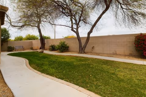 Outdoor garden area with a curved concrete pathway, green grass, several trees, bushes, and a bench near a beige brick wall under a cloudy sky.