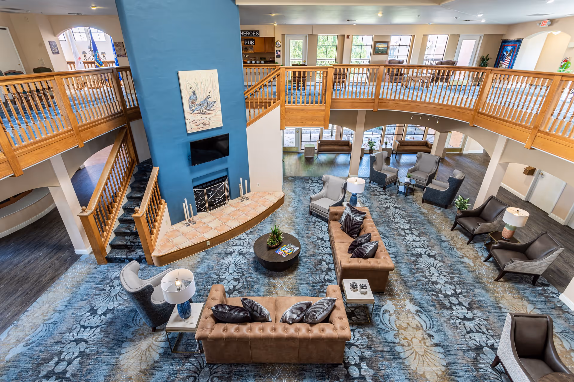 Spacious senior living facility common area with a high ceiling and a large blue fireplace in the center. The room features a patterned blue carpet, two brown tufted sofas with decorative pillows, several armchairs, side tables with lamps, and a wooden railing balcony overlooking the space. Large windows allow natural light to fill the room.