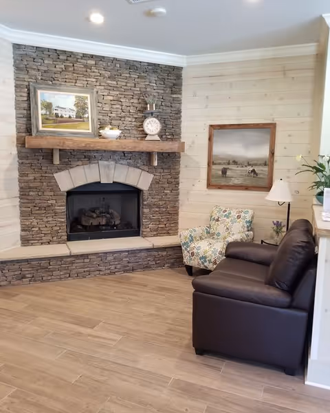 A cozy living room area featuring a stone fireplace with a wooden mantel. On the mantel are decorative items including a clock and a small plant. Above the mantel hangs a framed painting of a house. To the right of the fireplace is a floral patterned armchair and a dark brown leather sofa. A framed painting of cows in a field hangs on the light wood-paneled wall. A floor lamp and some flowers are also visible.