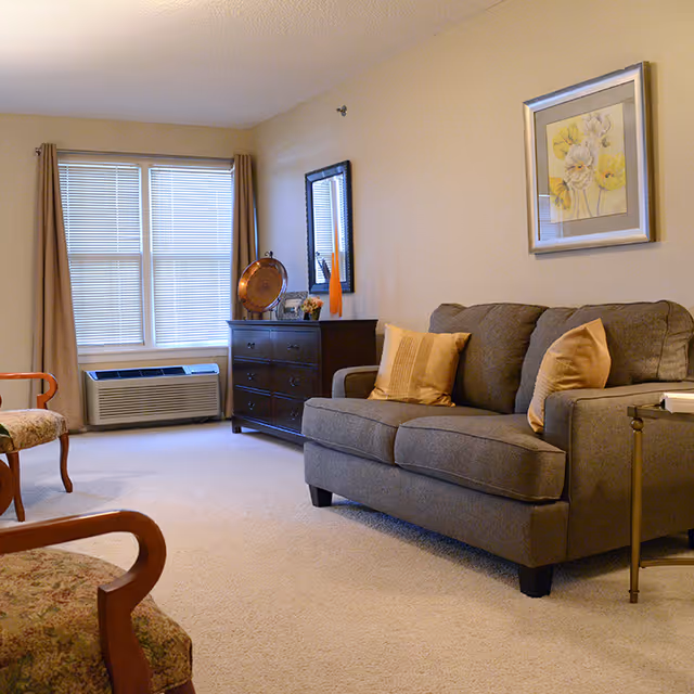 A cozy living room with a gray sofa adorned with two beige pillows, a wooden dresser with decorative items including a round copper tray and a tall orange vase, a framed floral painting on the wall, two upholstered chairs with wooden arms, beige carpet, and a window with closed blinds and beige curtains.