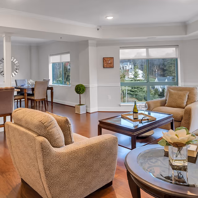 Open, well-lit living room and dining area with upholstered armchairs, a coffee table with a wine bottle and glass, and large windows showing trees outside.